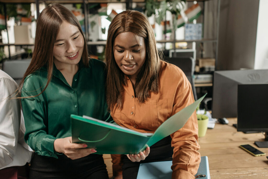 Updated Homepage: Two diverse women collaborate, reviewing documents in an office setting. Teamwork and updated homepage showcase.