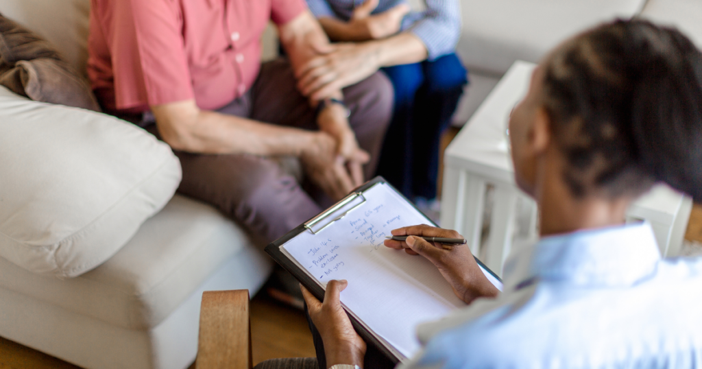 Counselor writes notes on a clipboard while group members sit on a sofa in a counseling session.