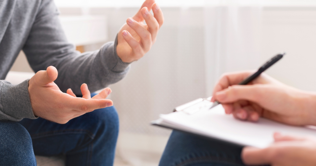 Person in a gray sweatshirt speaks with hand gestures while another person takes notes on a clipboard, suggesting a counseling session.
