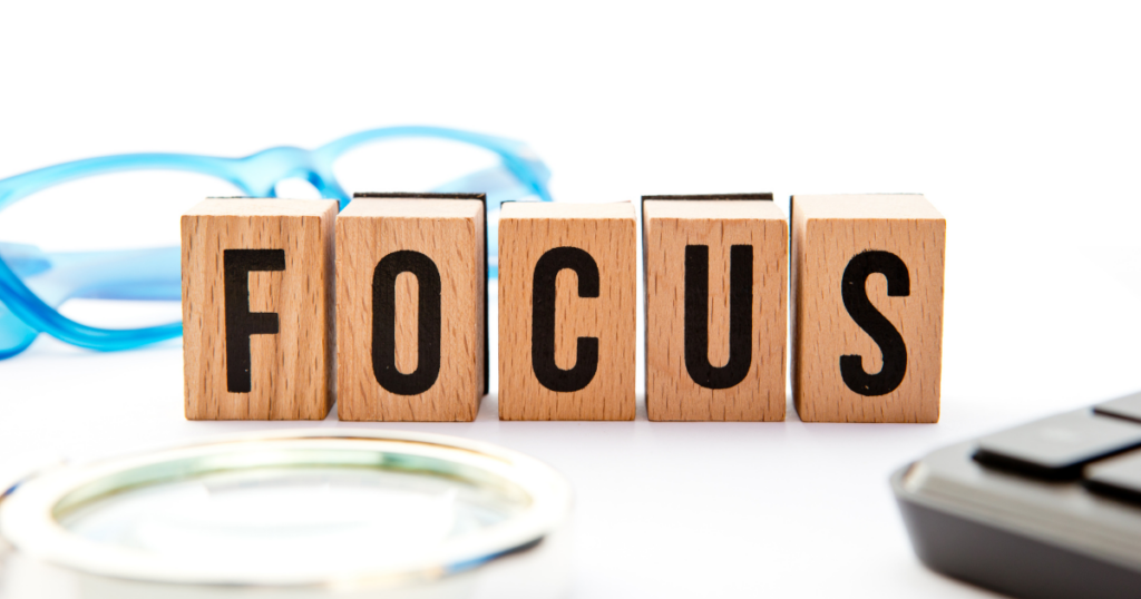 Wooden blocks spell 'FOCUS' on a white surface, with blue eyeglasses and a magnifying glass nearby.