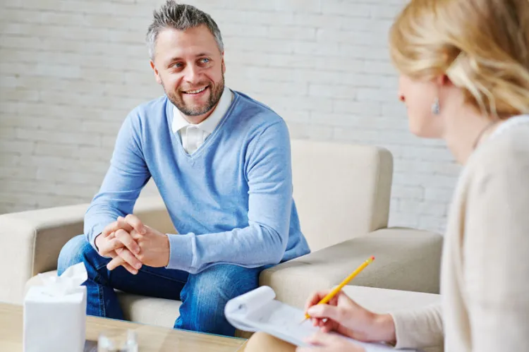 Man in a blue sweater speaks with a woman who is taking notes during a counseling session in a living room setting.