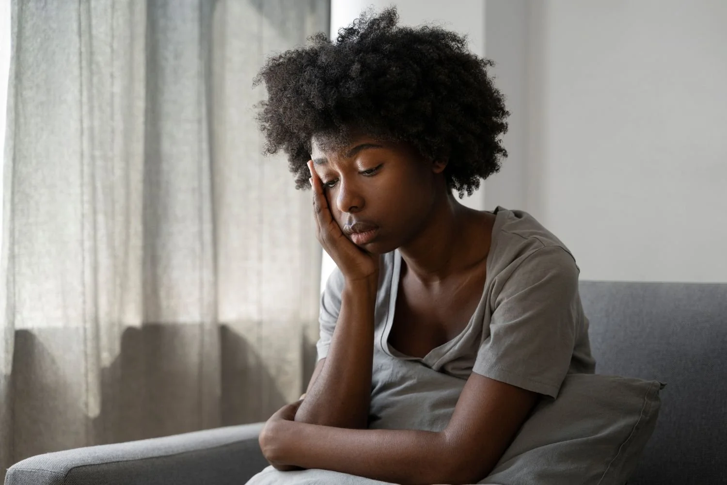 Woman sitting on a gray sofa with her head resting on her hand, looking sad or fatigued by the window.