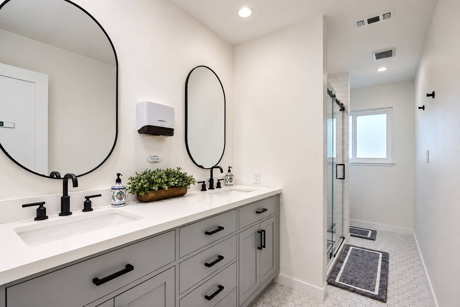 Double-sink bathroom vanity with gray cabinets, black faucets, and two oval mirrors above a white countertop and sinks.