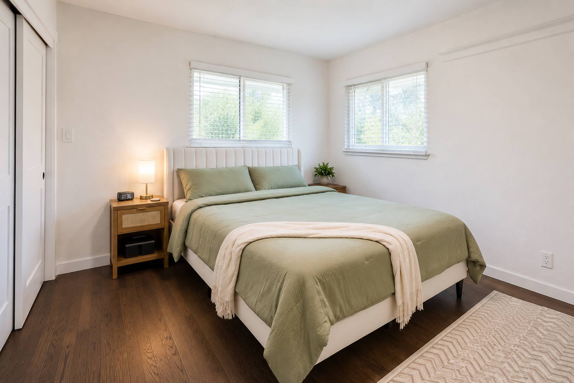 Bright bedroom with a green-olive quilted bed, white upholstered headboard, two matching nightstands, and two windows with blinds letting in light.
