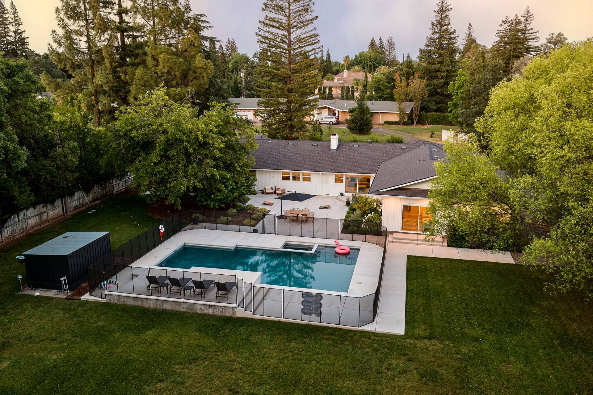 Aerial view of a backyard with a rectangular pool, glass fencing, and outdoor seating on a concrete patio surrounded by trees and a lawn.