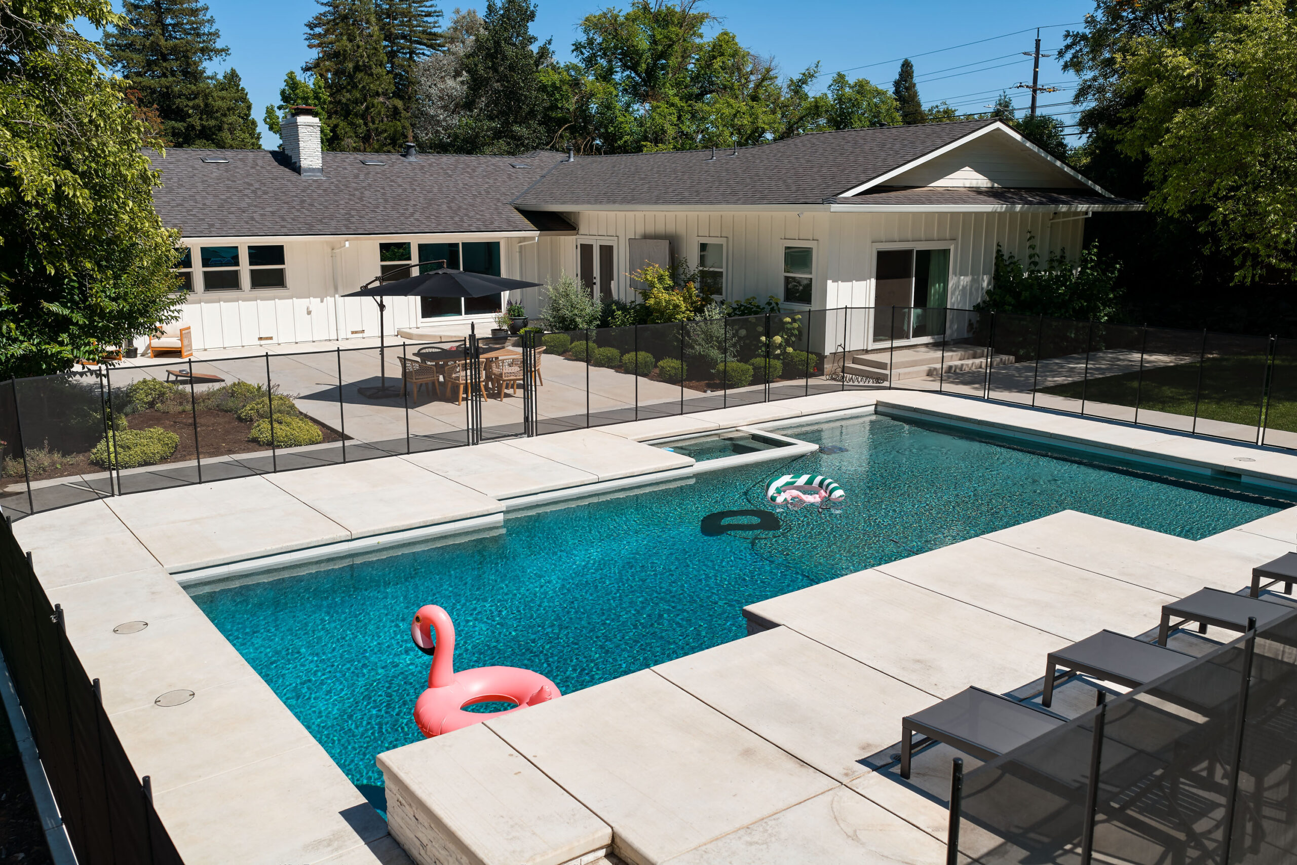 Poolside backyard with a rectangular pool, pink flamingo float, beige concrete deck, glass privacy fence, and white house with a dark roof in the background.