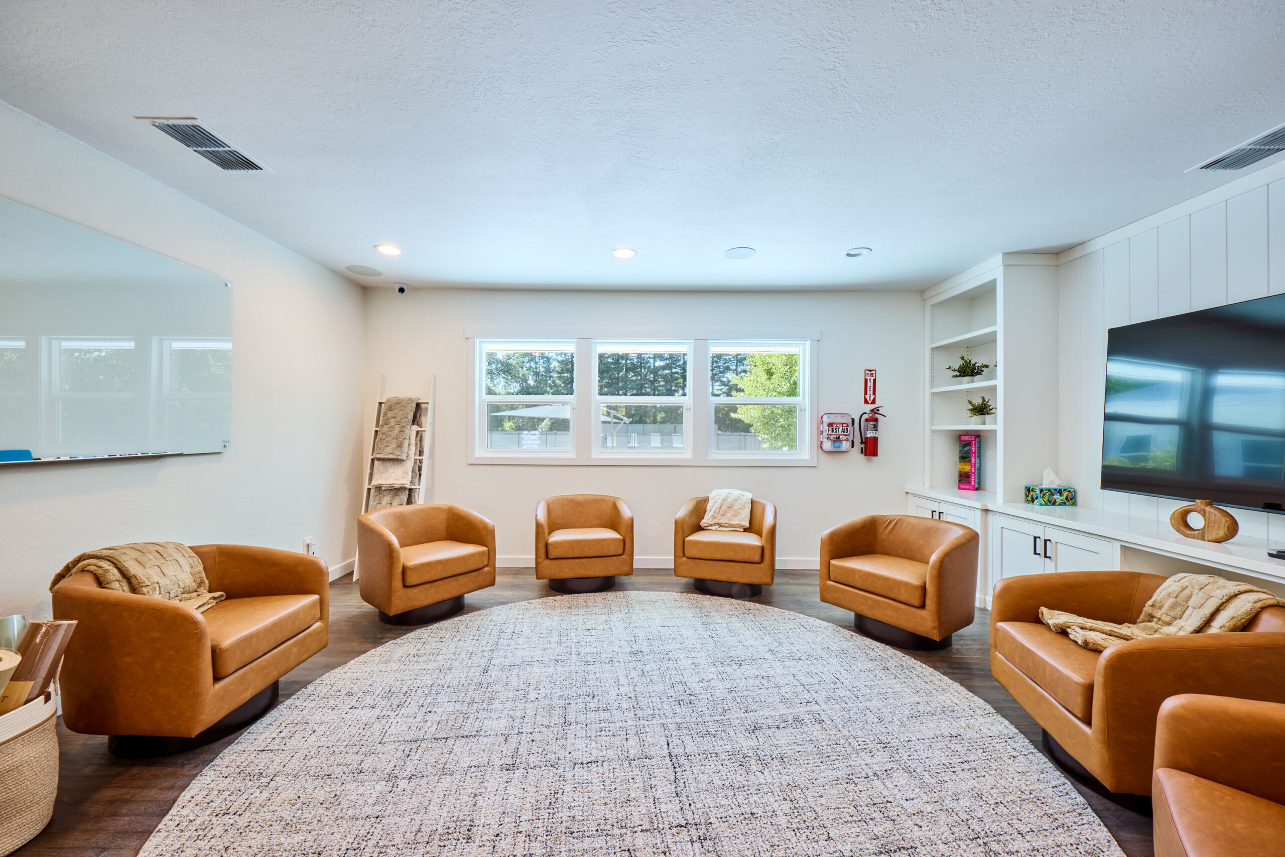 Bright living room lounge with six tan leather armchairs arranged around a circular rug, white built-in shelves, and a large wall-mounted TV to the right.