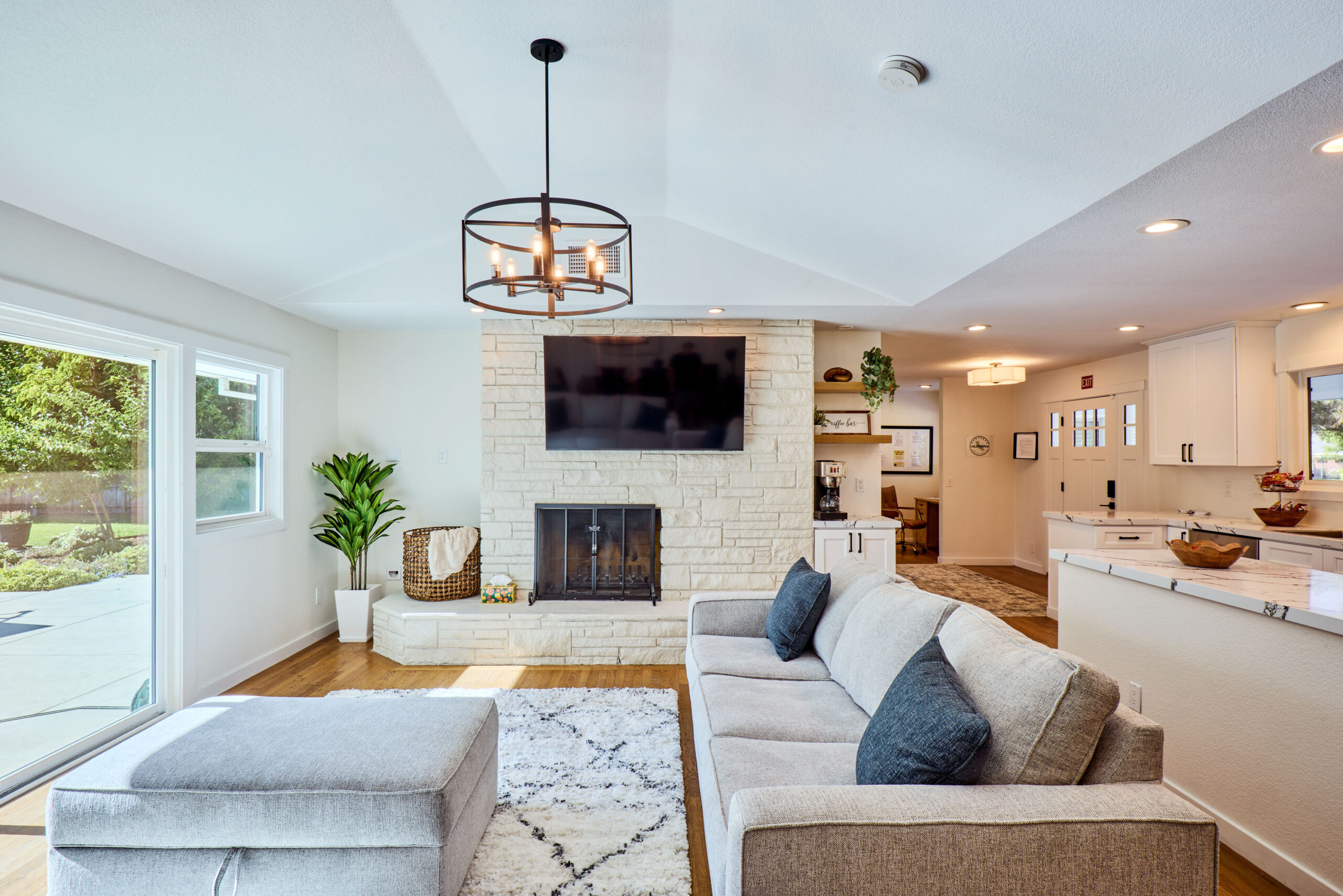 Sunlit living room with a stone fireplace, wall-mounted TV, gray sectional sofa, and a circular metal chandelier above the seating area.