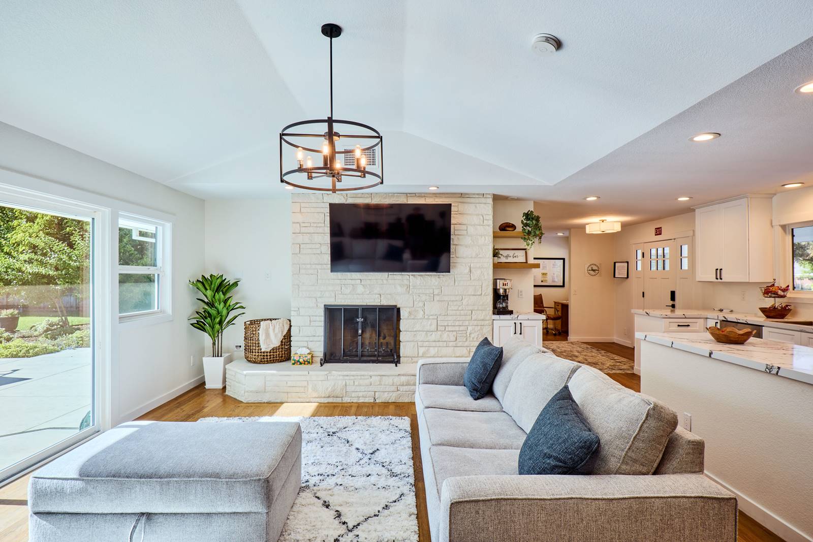 Bright open living room with a white stone fireplace, wall-mounted TV, and a gray sectional facing the kitchen area.