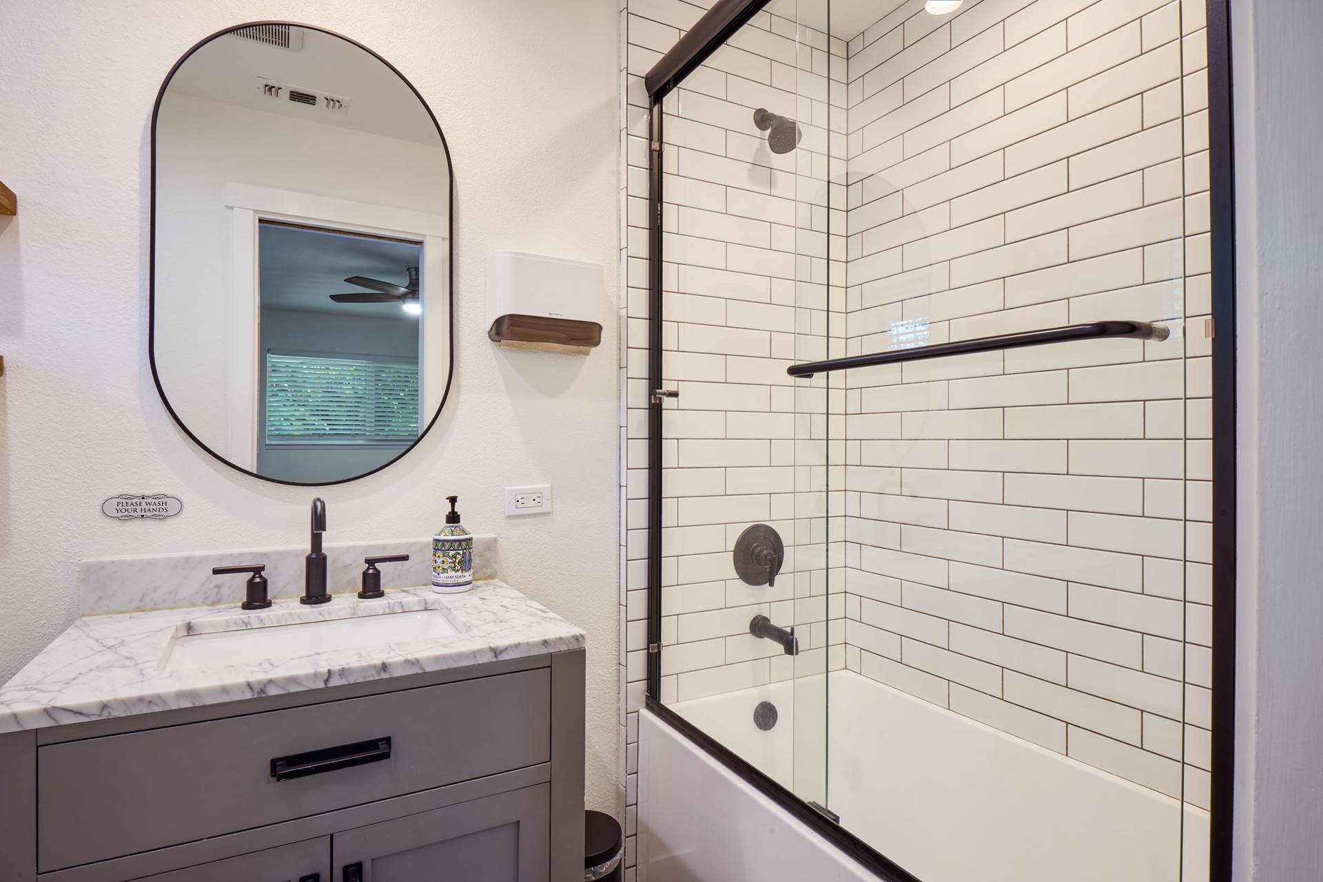 Gray bathroom vanity with marble countertop, oval mirror, and black fixtures; soap dispenser nearby.