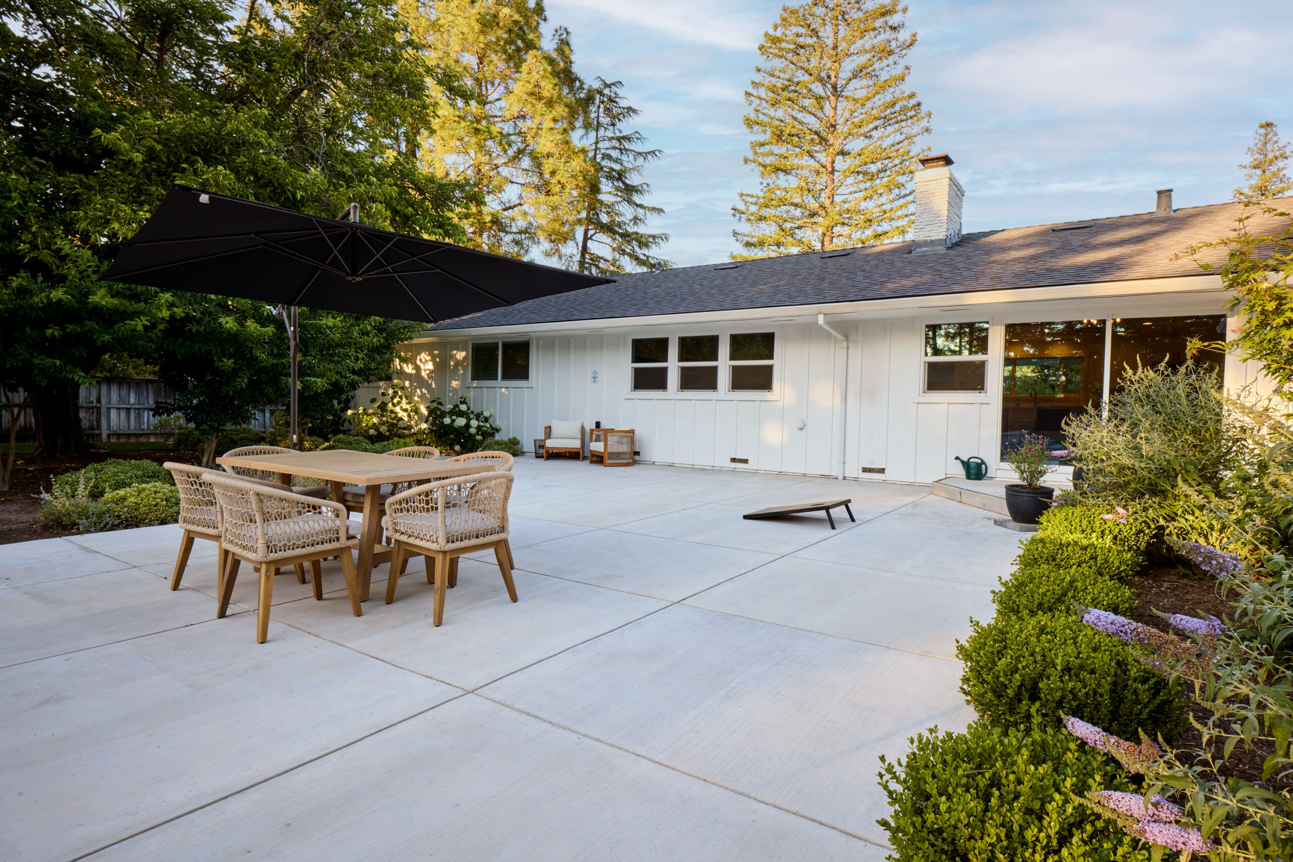 Backyard patio with a wooden dining table and woven chairs under a large black umbrella, in front of a white house.
