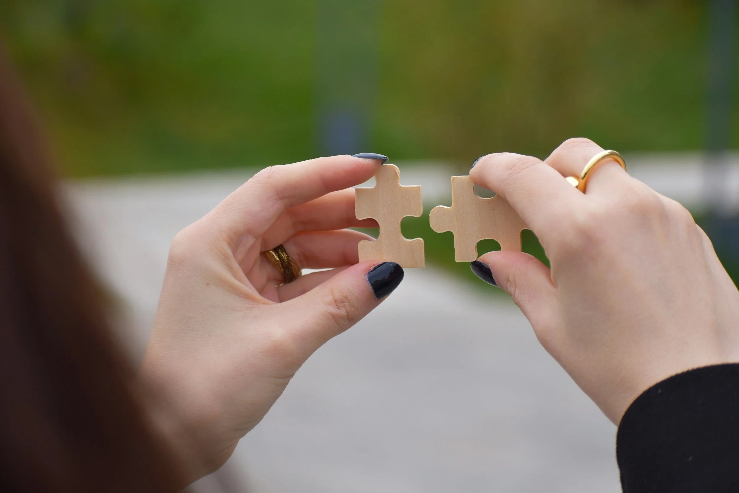 Two hands hold and interlock two wooden puzzle pieces against a blurred outdoor background, symbolizing connection or partnership with ringed fingers visible.