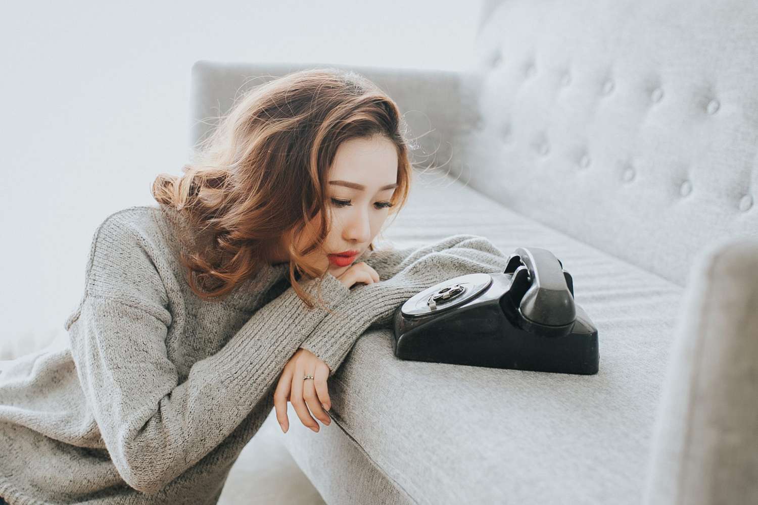 Woman in a gray sweater rests her head on a sofa arm, looking at a vintage black rotary phone on a gray couch.