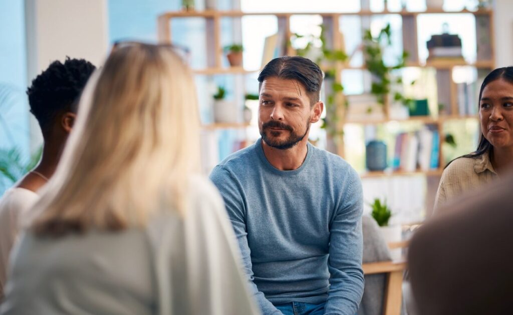 Man with a blue sweater listens during a group discussion in a bright open-office setting.