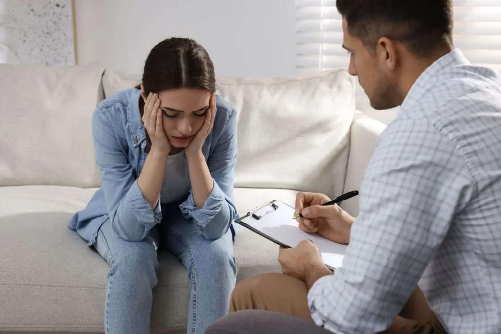 Woman sits on a sofa holding her head in distress while a man beside her takes notes on a clipboard with a pen.