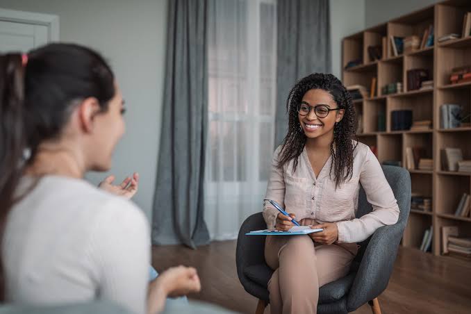 Therapist and client converse in a cozy room; therapist takes notes in a notepad and smiles at the client across from her.